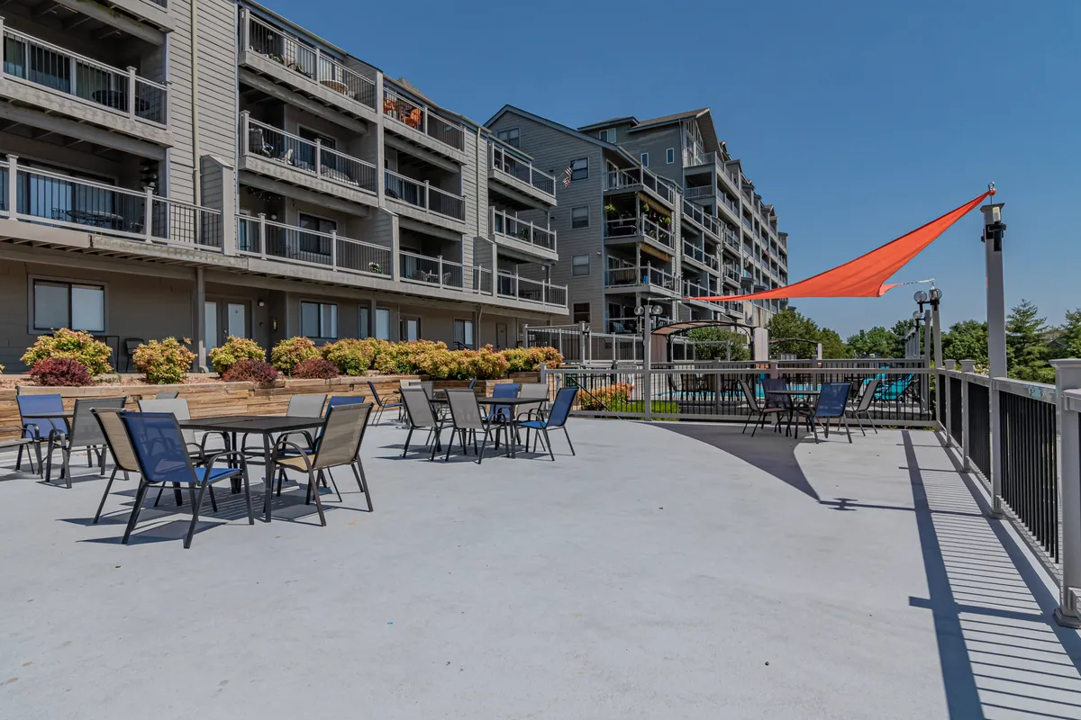 The image shows an outdoor seating area with tables and chairs under a bright orange shade sail, surrounded by colorful plants and facing the building's facade. The setting is lively and communal, perfect for socializing, casual meals, or relaxing outdoors with friends and couples.