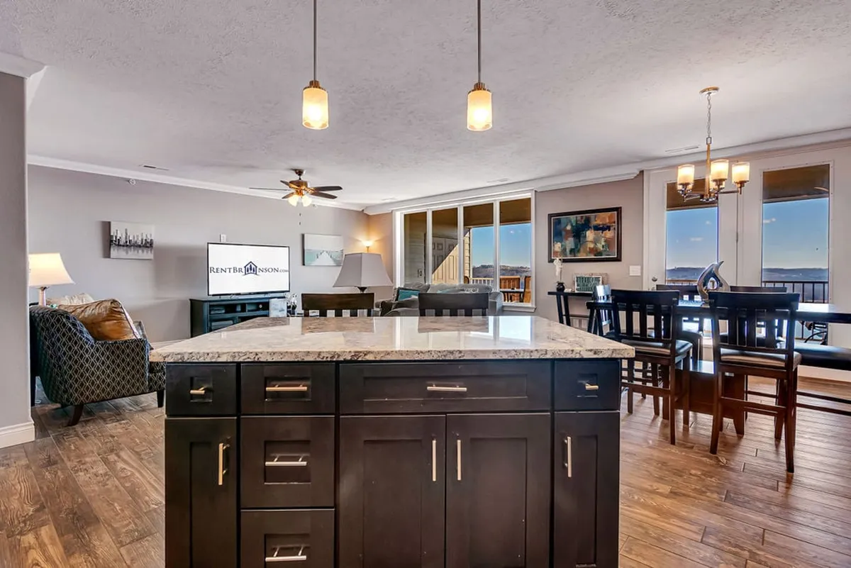 This image showcases an inviting, open-concept living area that flows seamlessly into the dining space and kitchen. Large windows fill the room with natural light and offer glimpses of lake views. The arrangement includes a plush sofa, dining table, and a kitchen island, illustrating a multi-functional hub perfect for gatherings and relaxation.