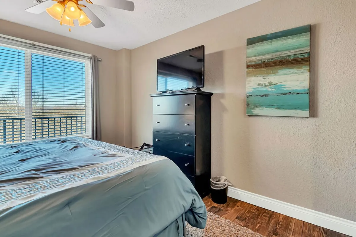 A peaceful bedroom featuring a bed dressed in blue bedding, a large window with adjustable blinds, a sleek black dresser topped with a television, and modern abstract wall art. The atmosphere is cozy and inviting.