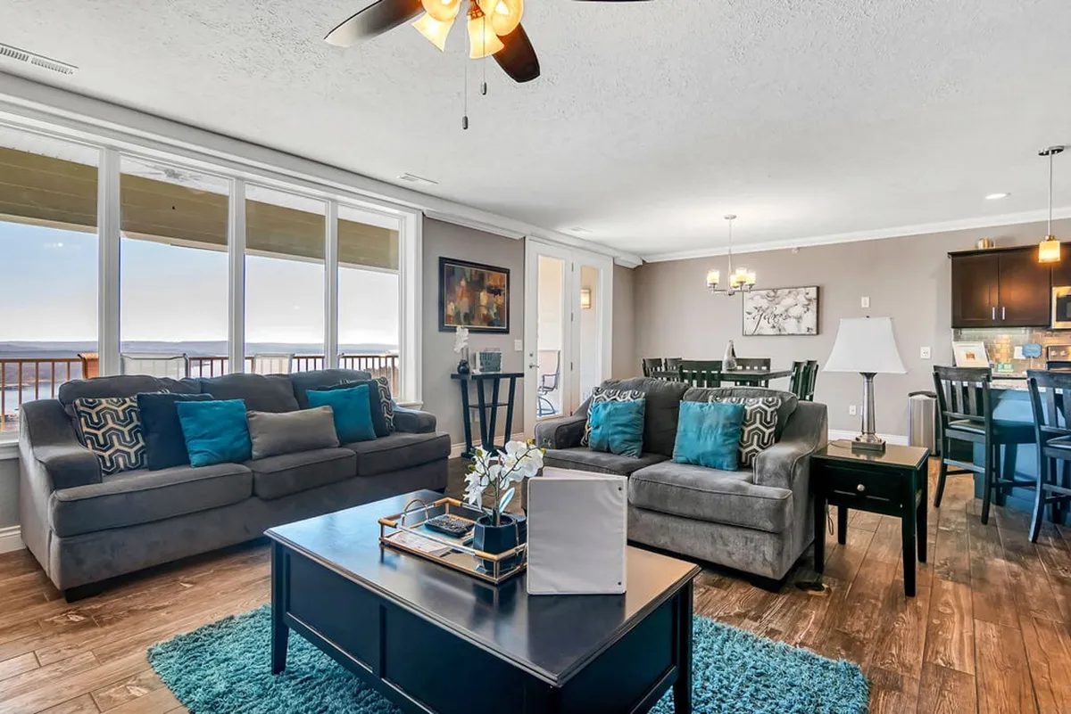The image showcases a beautifully decorated living area with plush, comfortable sofas arranged around a chic coffee table. Expansive windows frame breathtaking lake views, inviting natural light and revealing the stunning Branson sunset. The space is open and welcoming, perfect for relaxing evenings, family gatherings, or simply soaking in the scenic vistas from the comfort of the condo.