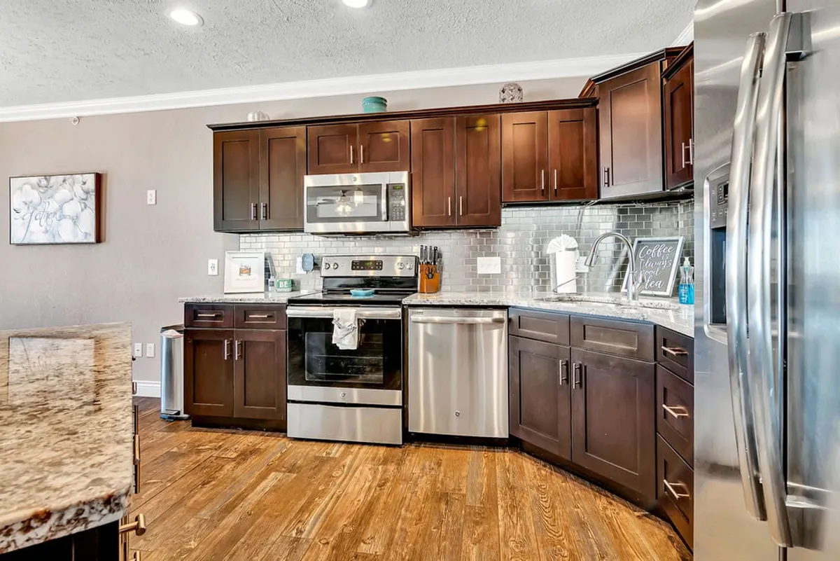 A view of the kitchen emphasizing sleek stainless steel appliances, dark wood cabinets, and a gleaming granite countertop. The backsplash tiles and open layout evoke a sense of warmth and welcoming, ideal for both everyday meals and special celebrations.