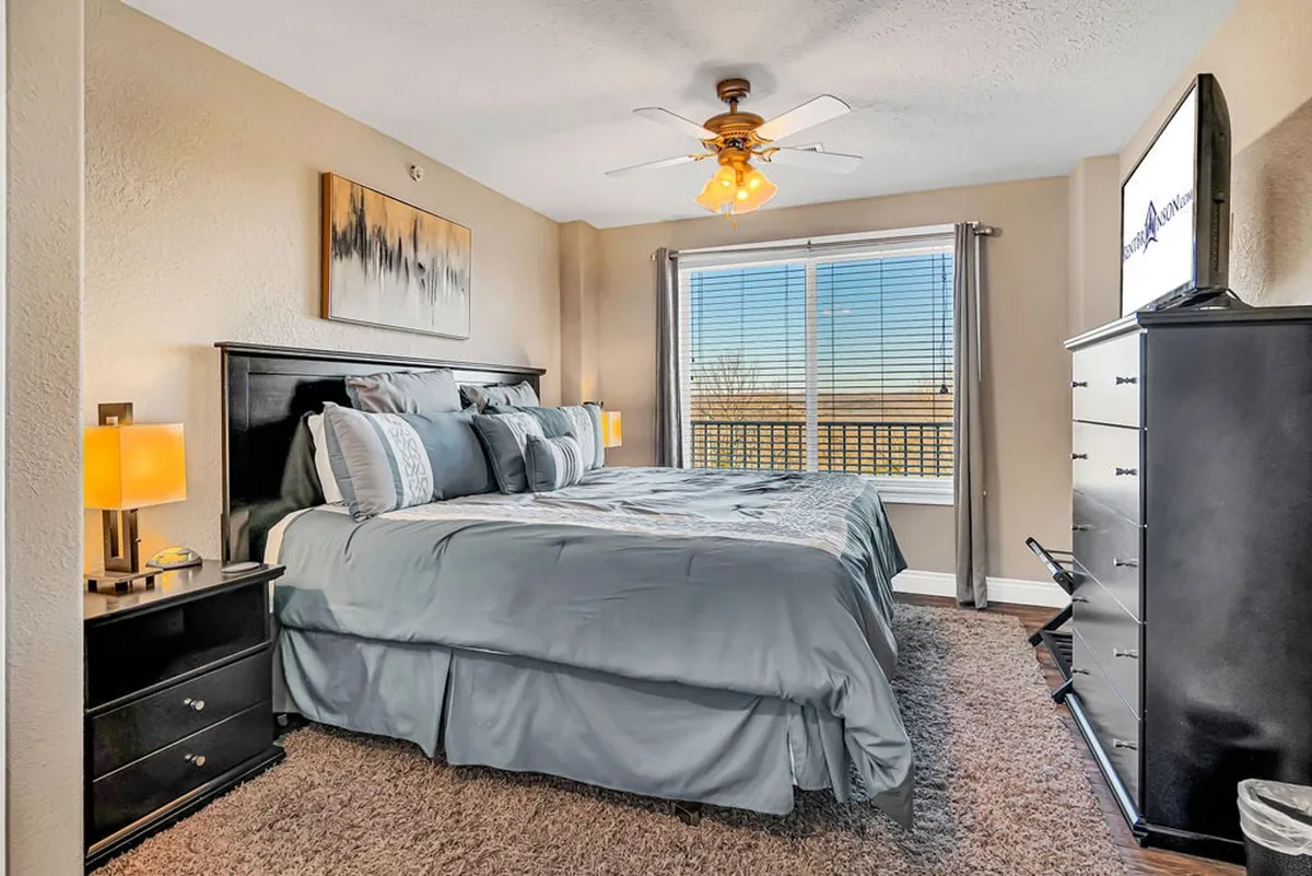 A bright and spacious bedroom with a large bed adorned with decorative pillows, flanked by nightstands with lamps. Natural light pours in through a window, and tasteful artwork hangs on the wall, creating a soothing, restful environment.
