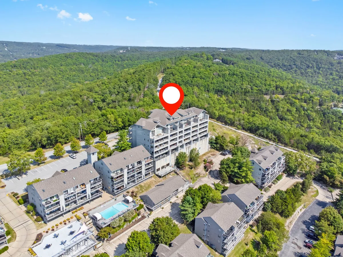 An exterior shot of the multi-story property building with the swimming pool in the foreground, surrounded by greenery and a visible parking area. The atmosphere is relaxed, showing ample guest parking, access to resort amenities, and the overall building layout.