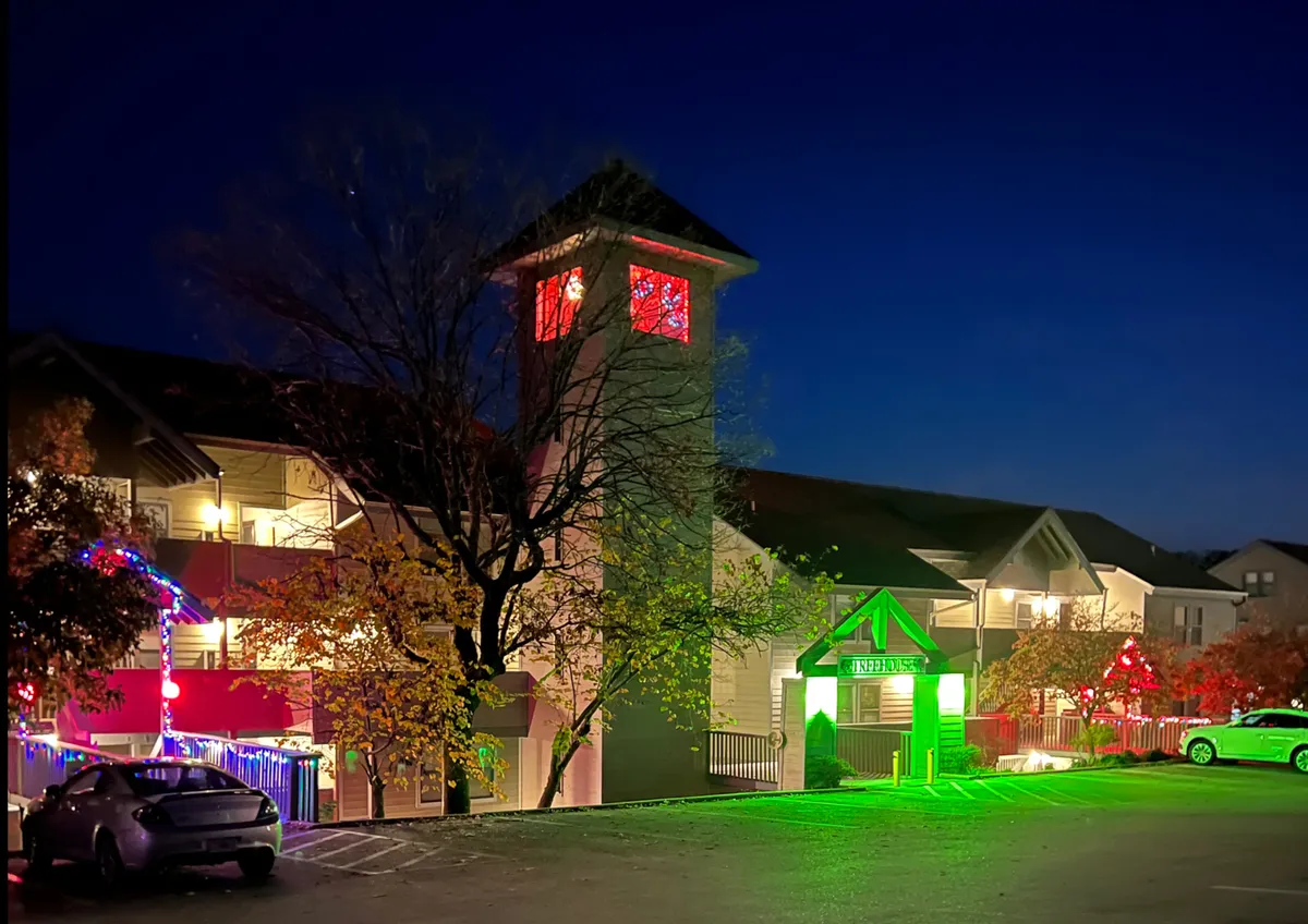 A festive exterior scene featuring a decorated tree in the foreground, an illuminated clock tower, colorful lights on surrounding buildings, and a glowing entrance sign. The image conveys a sense of celebration, evening energy, and shared experiences in a lively neighborhood environment.